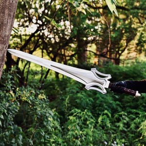 May include: A silver-colored sword with a pointed blade and a complex, multi-layered crossguard. The handle is wrapped in dark brown material with a light-colored pommel at the end, held horizontally against a tree trunk in a wooded area.