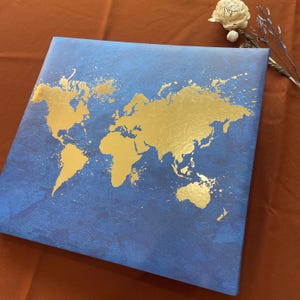May include: A square canvas wall art piece featuring a deep blue background with a textured, mottled finish. A gold-colored world map is printed across the center, characterized by a splattered, paint-like effect along the continental coastlines.