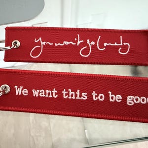 May include: Two rectangular red fabric keychains with silver metal rings. The top keychain features white cursive embroidery reading You won't go lonely. The bottom keychain features white block letter embroidery reading We want this to be good.