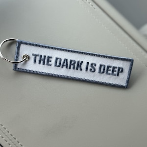 May include: A rectangular white fabric keychain with a dark gray embroidered border and the text THE DARK IS DEEP stitched in dark gray thread. A silver metal split ring is attached to the left side through a reinforced metal eyelet.