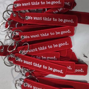 Puede incluir: Una colección de llaveros rectangulares de tela roja con anillas de metal plateado. Cada llavero está bordado con el texto en blanco "We want this to be good.". Los artículos están apilados, mostrando el diseño y las letras uniformes.