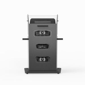 May include: A dark gray vertical kiosk with a rectangular frame, featuring three recessed horizontal compartments with integrated LED light arrays, side handles, and a clear acrylic top panel, standing on a wide, flat base.