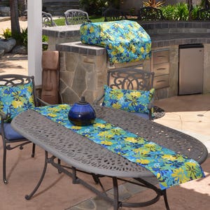 May include: An outdoor dining set featuring a dark gray metal table and chairs with blue, yellow, and green floral cushions and a matching table runner. A dark blue ceramic oil lamp sits on the table, and a matching floral cover is on the grill.