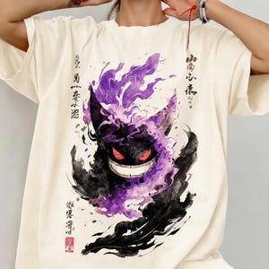 May include: An off-white short-sleeve t-shirt featuring a large graphic print of a dark purple and black creature with glowing red eyes and a wide grin, surrounded by purple smoke-like wisps and vertical columns of black Japanese calligraphy text.