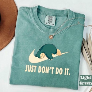 May include: A light green crew neck t-shirt featuring a graphic of a sleeping Snorlax character resting on a cream-coloured swoosh logo. Below the graphic, the text reads JUST DON'T DO IT. The shirt has a Comfort Colors brand label inside the collar.