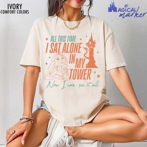 以下が含まれることがあります： 「ALL THIS TIME I SAT ALONE IN MY TOWER Now I can see it all.」というテキストがプリントされたアイボリーの半袖Tシャツ。デザインには、長い髪のキャラクターと高い塔のオレンジ色のイラストが描かれています。