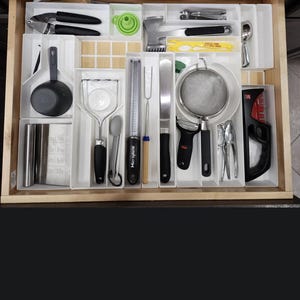 May include: A wooden kitchen drawer organizer with multiple white plastic dividers containing various cooking utensils including a metal meat mallet, a Microplane grater, a potato masher, a mesh strainer, a vegetable peeler, and a black measuring cup.