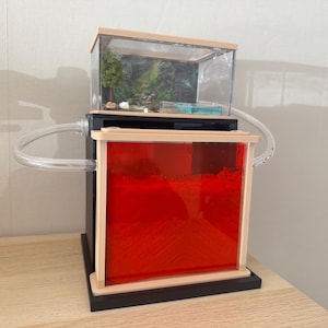 Op de afbeelding: Een mierenboerderij met twee niveaus, voorzien van een zwarte basis en frame. Het bovenste niveau is een doorzichtige behuizing met een miniatuur bosdiorama met een boom en rotsen. Het onderste niveau is een vierkante bak met rode panelen.