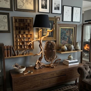 Puede incluir: Una lámpara de mesa con base de madera flotante natural y pantalla de tela negra. Un reloj circular de rodaja de madera con manecillas finas cuelga de la rama. La lámpara se apoya sobre una consola de madera larga con cajones y superficie lisa.