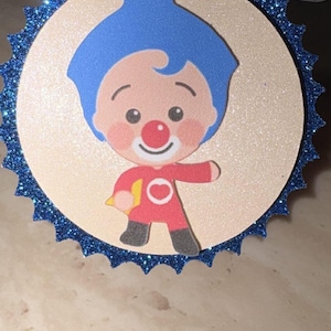 May include: A circular decorative topper featuring a cartoon character with blue hair, a red nose, and a red outfit with a white circle emblem. The character is centered on a light beige disc, which is framed by a dark blue glittery scalloped border.