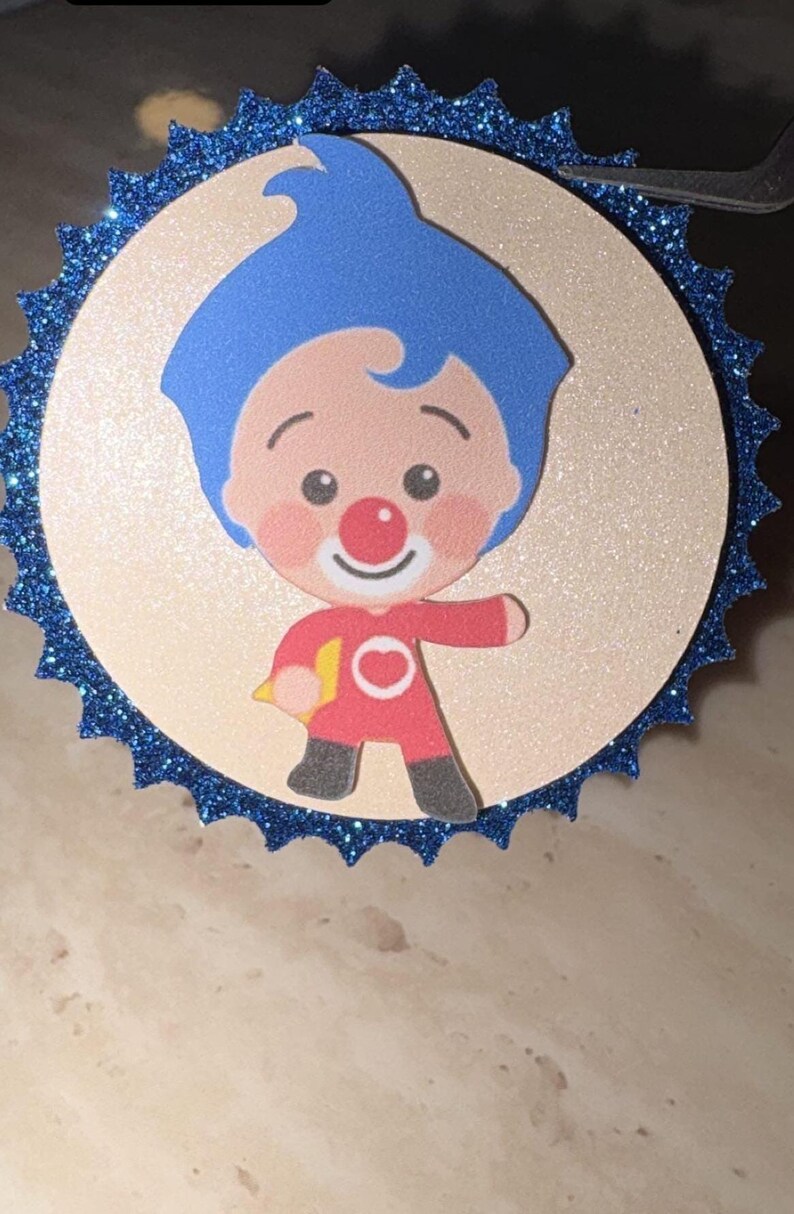 May include: A circular decorative topper featuring a cartoon character with blue hair, a red nose, and a red outfit with a white circle emblem. The character is centered on a light beige disc, which is framed by a dark blue glittery scalloped border.