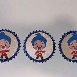 May include: Three circular cupcake toppers featuring a cartoon character with blue hair and a red outfit. Each topper has a beige center with the character illustration and a scalloped border made of dark blue glittery material.