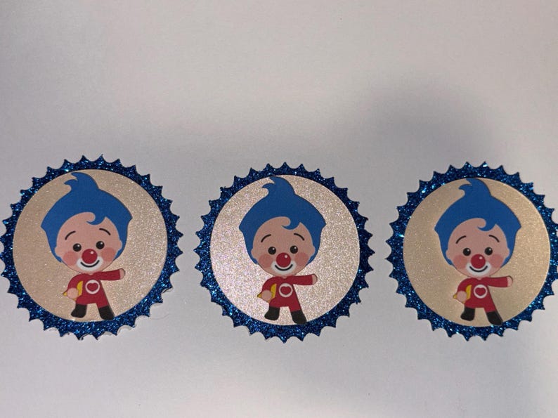 May include: Three circular cupcake toppers featuring a cartoon character with blue hair and a red outfit. Each topper has a beige center with the character illustration and a scalloped border made of dark blue glittery material.