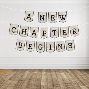 May include: A banner with the words "A NEW CHAPTER BEGINS" strung across a white wall. Each pennant is a light beige color with black text and a black border. The background is a white wall and a wooden floor.
