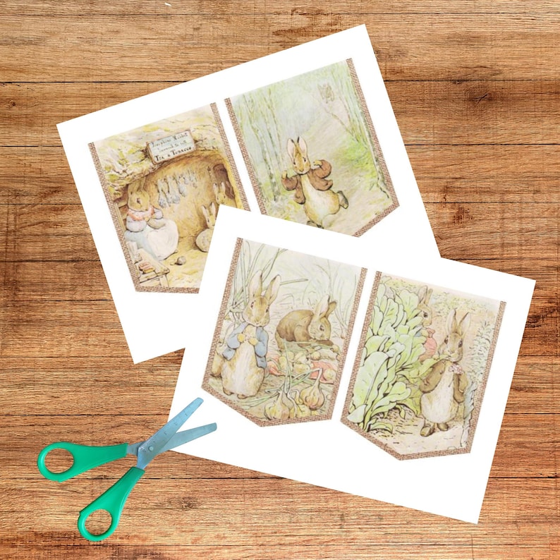 May include: Three illustrated paper banners featuring Peter Rabbit scenes, with a gold glitter border. The banners depict rabbits in various settings, including a garden and a burrow. A pair of green-handled scissors is in the foreground.