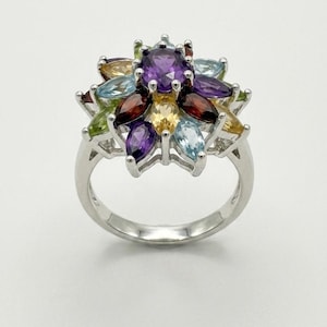 May include: A silver ring featuring a central round purple amethyst surrounded by a cluster of marquise-cut gemstones in various colors, including light blue, yellow, deep red, and green, arranged in a floral pattern on a polished silver band.