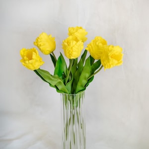 May include: A bouquet of six vibrant yellow parrot tulips with ruffled petals and green stems arranged in a clear, vertically ribbed glass vase. The flowers feature distinct, wavy petal edges and are displayed against a neutral background.