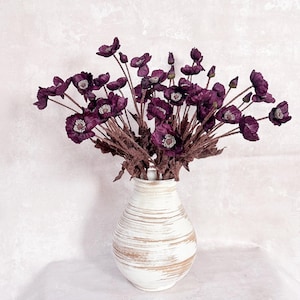 Peut inclure: Un bouquet de fleurs de pavot violet foncé avec des tiges et des centres bruns texturés, disposé dans un vase en céramique blanche présentant des motifs de coups de pinceau bruns horizontaux et usés sur le corps arrondi du récipient.