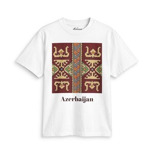 Könnte beinhalten: Ein weißes kurzärmeliges T-Shirt mit einer zentralen rechteckigen Grafik auf dunkelrotem Hintergrund, komplexen geometrischen Mustern in Gold, Grün und Blau sowie dem Wort Azerbaijan in dunkelbrauner Schrift unter dem Design.