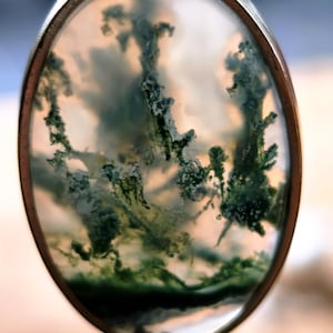 May include: An oval pendant featuring a translucent moss agate gemstone set in a smooth silver-toned metal frame. The stone displays intricate, branching green inclusions that resemble forest foliage against a cloudy, off-white background.