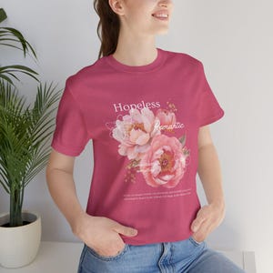 Puede incluir: Una camiseta de manga corta de color rosa oscuro con un gráfico central de peonías rosas y flores dibujadas con líneas blancas. El texto dice Hopeless Romantic, Romantizing everything, y un párrafo de texto pequeño debajo del diseño.