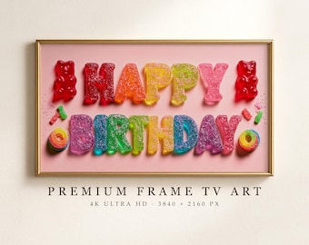 Puede incluir: Un cuadro rectangular con marco dorado que muestra el texto HAPPY BIRTHDAY formado por letras de gominola de colores recubiertas de azúcar sobre un fondo rosa, flanqueado por ositos de goma rojos y pequeños anillos de goma.