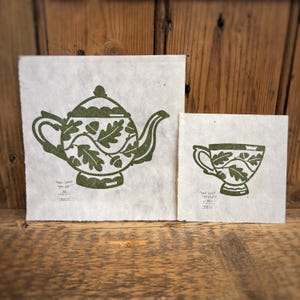 May include: Two green hand-printed illustrations on paper. The larger illustration is of a teapot with an oak leaf design. The smaller illustration is of a teacup with an oak leaf design. Both illustrations have the text "One Leaf Teapot" written below them.