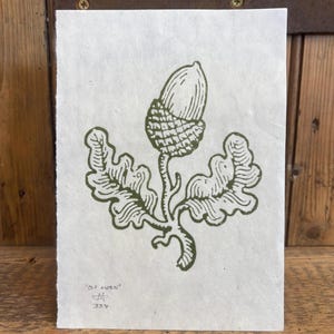 May include: A green ink print of an acorn and oak leaves on textured white paper. The artwork includes the text "old acorn" and artist's initials. The print is set against a wooden background.