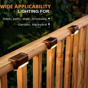 May include: Three brown solar-powered deck lights mounted on a wooden railing. The text reads: WIDE APPLICABILITY LIGHTING FOR Deck, path, stair, Driveway Garden, backyard. The lights emit a warm glow downward along the wooden posts.
