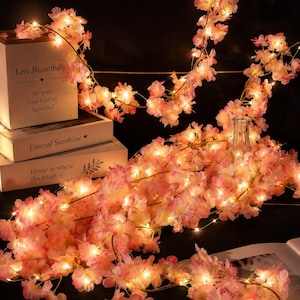 Könnte beinhalten: Eine Lichterkette mit künstlichen Kirschblüten in Rosa- und Weißtönen, beleuchtet durch kleine warmweiße LED-Lichter. Die Blumengirlande ist über drei gestapelte weiße rechteckige Schachteln mit Text wie Live Beautifully drapiert.