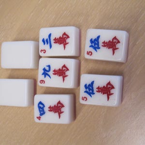 May include: A collection of white Mahjong tiles with red and blue symbols and numbers. The tiles are arranged on a light brown surface. The numbers 3, 4, 5, and 9 are visible on the tiles.