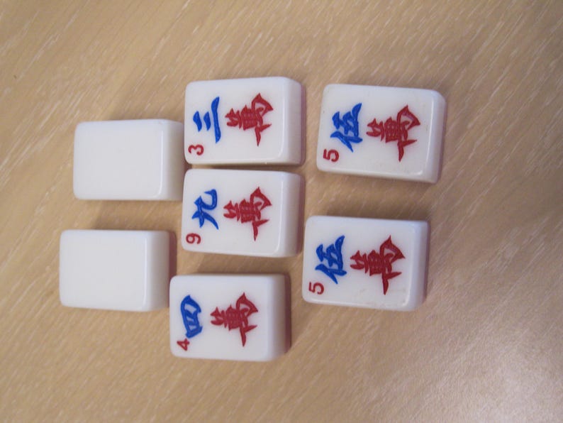 May include: A collection of white Mahjong tiles with red and blue symbols and numbers. The tiles are arranged on a light brown surface. The numbers 3, 4, 5, and 9 are visible on the tiles.
