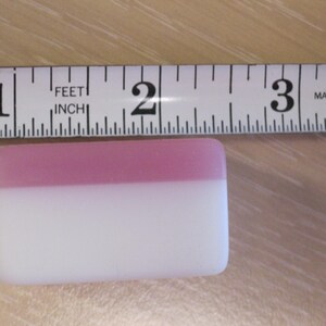 May include: A rectangular, two-toned object, approximately 2 inches long, with a pink top and white bottom. A measuring tape is placed above the object, indicating its length. The tape is marked in inches and feet.