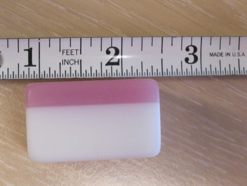 May include: A rectangular, two-toned object, approximately 2 inches long, with a pink top and white bottom. A measuring tape is placed above the object, indicating its length. The tape is marked in inches and feet.