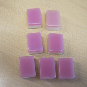 May include: Seven rectangular mahjong tiles with a pink top and white bottom are arranged on a light brown wooden surface. The tiles are smooth and have a glossy finish, suggesting they are made of a durable material.
