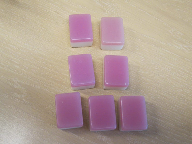 May include: Seven rectangular mahjong tiles with a pink top and white bottom are arranged on a light brown wooden surface. The tiles are smooth and have a glossy finish, suggesting they are made of a durable material.