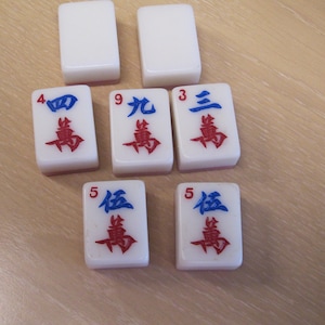 May include: White Mahjong tiles with blue and red symbols. The tiles are arranged on a light brown surface. The tiles display the numbers 3, 4, 5, and 9 in Chinese characters.