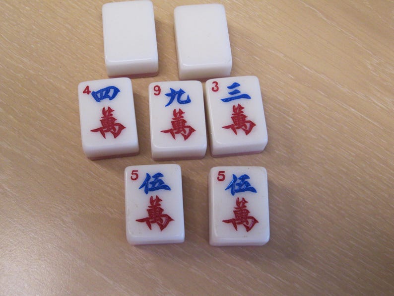 May include: White Mahjong tiles with blue and red symbols. The tiles are arranged on a light brown surface. The tiles display the numbers 3, 4, 5, and 9 in Chinese characters.