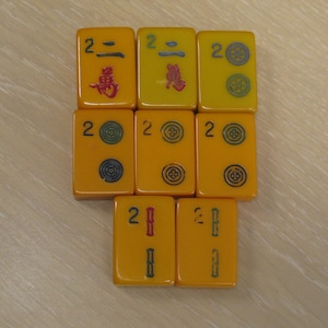 May include: Eight rectangular yellow Mahjong tiles arranged in a grid. The tiles feature the number 2, Chinese characters, and circular or bamboo-style symbols in blue, red, and green ink. Each tile has a smooth, glossy finish.