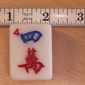 May include: A white Mahjong tile with the number 4 in red and a blue symbol. The tile is approximately 1 inch wide, as measured by a ruler in the background. The tile has a red symbol at the bottom.
