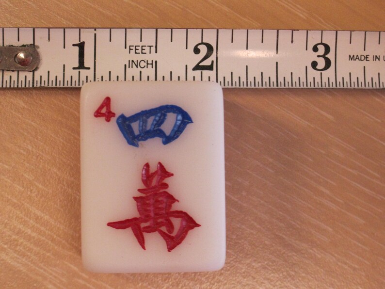 May include: A white Mahjong tile with the number 4 in red and a blue symbol. The tile is approximately 1 inch wide, as measured by a ruler in the background. The tile has a red symbol at the bottom.