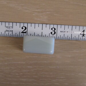 K&ouml;nnte beinhalten: Ein kleiner, rechteckiger, hellblauer Glasfliese, etwa 3,8 cm lang, wird auf einer Holzoberfl&auml;che gezeigt. Die Fliese hat abgerundete Kanten und ein subtiles, geometrisches Design. Ein Ma&szlig;band ist im Hintergrund.