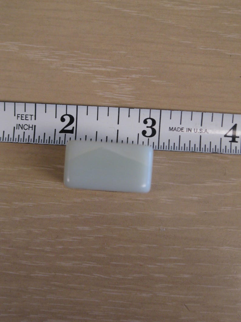 K&ouml;nnte beinhalten: Ein kleiner, rechteckiger, hellblauer Glasfliese, etwa 3,8 cm lang, wird auf einer Holzoberfl&auml;che gezeigt. Die Fliese hat abgerundete Kanten und ein subtiles, geometrisches Design. Ein Ma&szlig;band ist im Hintergrund.