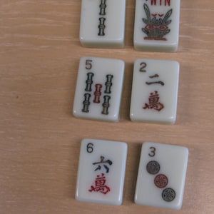 K&ouml;nnte beinhalten: Eine Sammlung elfenbeinfarbener Mahjong-Steine. Die Steine zeigen verschiedene Symbole und Zahlen in Rot und Schwarz, darunter Bambus, Kreise und chinesische Schriftzeichen. Ein Stein zeigt "4 WIN" mit einem floralen Design.