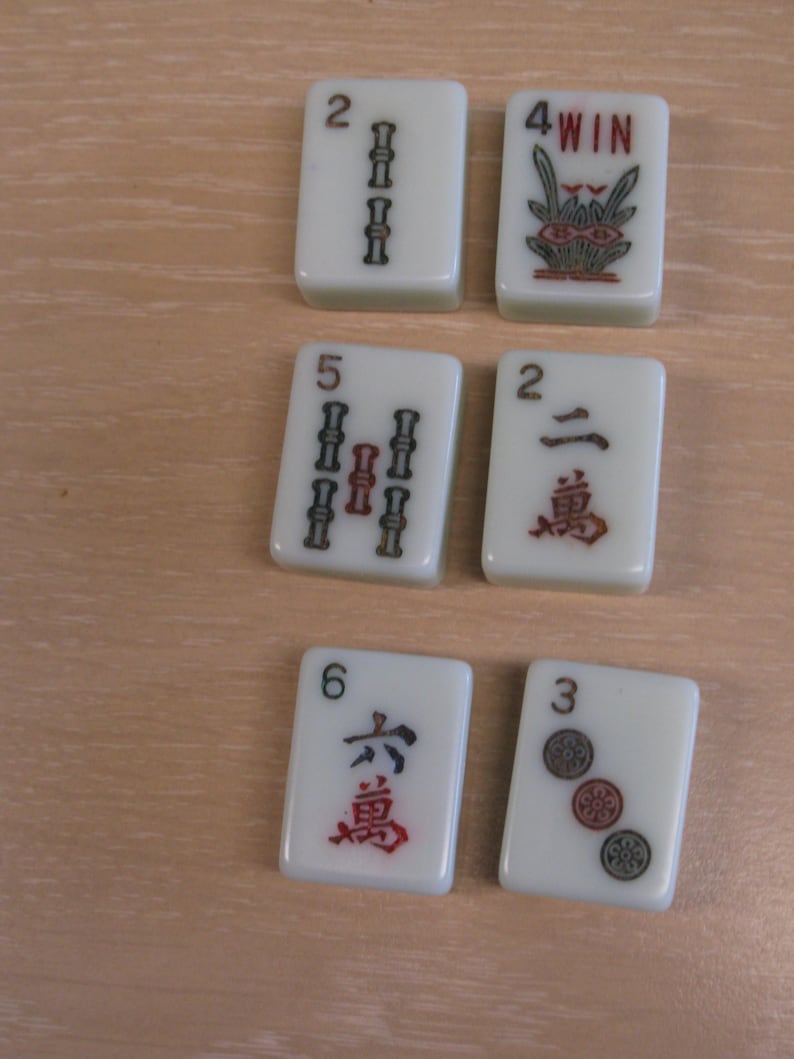 K&ouml;nnte beinhalten: Eine Sammlung elfenbeinfarbener Mahjong-Steine. Die Steine zeigen verschiedene Symbole und Zahlen in Rot und Schwarz, darunter Bambus, Kreise und chinesische Schriftzeichen. Ein Stein zeigt "4 WIN" mit einem floralen Design.