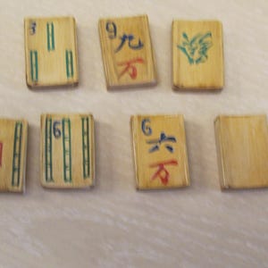 K&ouml;nnte beinhalten: Sieben Vintage-Mahjong-Steine mit bambusartiger Textur. Die Steine zeigen verschiedene chinesische Schriftzeichen und Symbole in roter, gr&uuml;ner und blauer Tinte. Die Steine sind auf einer hellen Oberfl&auml;che angeordnet.