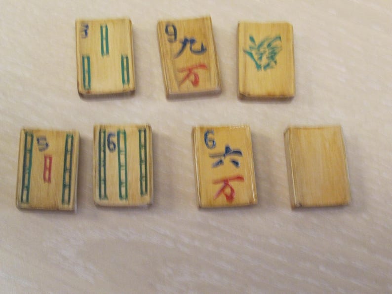 K&ouml;nnte beinhalten: Sieben Vintage-Mahjong-Steine mit bambusartiger Textur. Die Steine zeigen verschiedene chinesische Schriftzeichen und Symbole in roter, gr&uuml;ner und blauer Tinte. Die Steine sind auf einer hellen Oberfl&auml;che angeordnet.