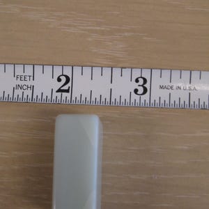 K&ouml;nnte beinhalten: Ein rechteckiger, hellgr&uuml;ner Gegenstand, etwa 6,35 cm lang, liegt in der N&auml;he eines Ma&szlig;bands. Das Objekt hat abgerundete Kanten und eine glatte Oberfl&auml;che. Das Ma&szlig;band ist in Zoll markiert und tr&auml;gt die Aufschrift "MADE IN U.S.A."