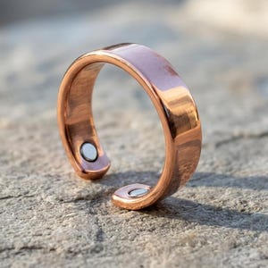 May include: A polished copper-colored open band ring featuring a smooth, metallic finish. The ring has a gap at the front and includes two small, circular silver-toned magnetic inserts embedded on the inner ends of the band.