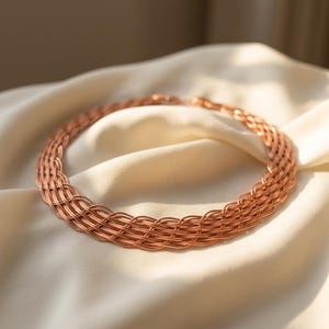 May include: A circular necklace constructed from multiple strands of copper-colored wire woven into a complex, repeating lattice pattern. The metallic piece features a consistent, interconnected design that forms a wide, flexible band structure.